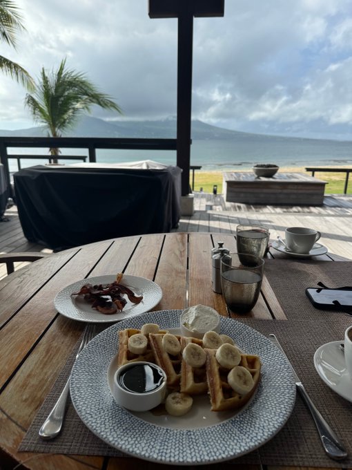 A breakfast spread of banana-topped waffles with syrup, bacon, and coffee is set on a wooden table overlooking a tropical ocean view, suggesting the poster is enjoying a leisurely morning break from the crypto markets.