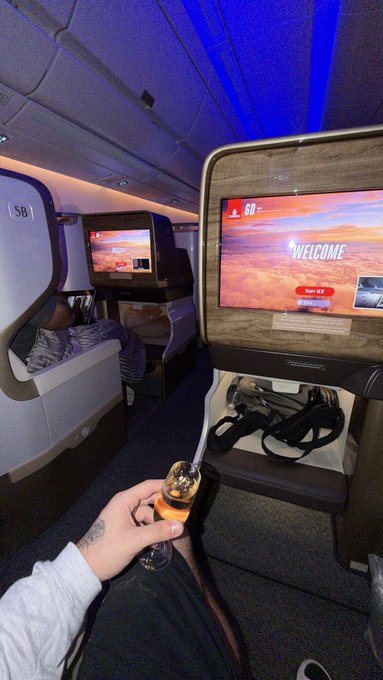 A hand holding a glass of champagne toasts in a business class cabin (seat 5B) with "WELCOME" screens and "60" anniversary branding visible, matching the post's celebratory "Back to Dubai" return trip.