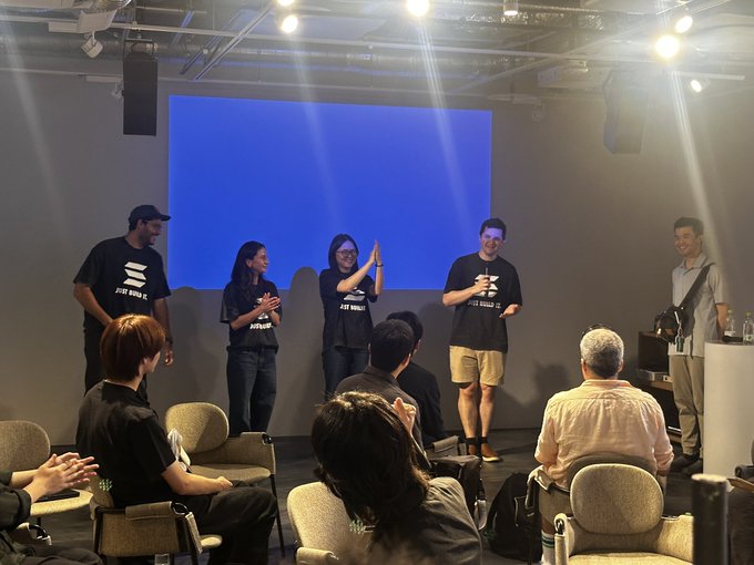 The image shows a group of people standing around, with some sitting down and others standing up. They are all facing a large screen that displays a blue background. The people appear to be engaged in an event or gathering, possibly a presentation or discussion. One person is holding a microphone, suggesting they might be speaking or leading the conversation.

There are several chairs visible in the scene, with some occupied and others empty. A few individuals can also be seen wearing backpacks, indicating that they may have brought their belongings to the event. The overall atmosphere seems to be one of collaboration and engagement among the participants.