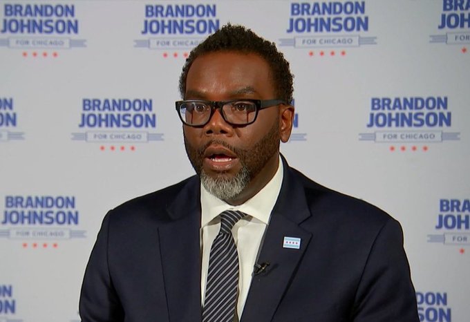 Brandon Johnson, wearing glasses, a navy suit and striped tie, speaks in front of a backdrop repeating 'BRANDON JOHNSON FOR CHICAGO'.
