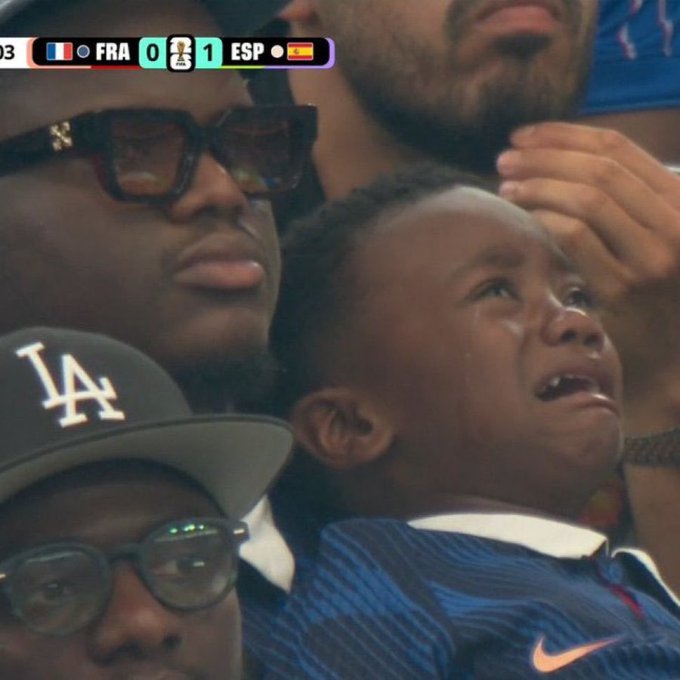 A young boy in a blue Nike shirt cries with tears streaming down his face while sitting among adults, one wearing an LA cap. A scoreboard graphic shows FRA 0, ESP 1 in the top left corner.