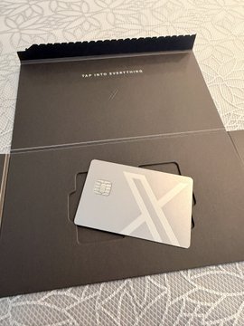 A metallic silver card with an 'X' logo and chip, displayed on a black presentation folder that reads 'TAP INTO EVERYTHING'.