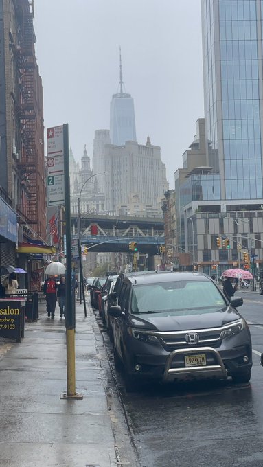 A rainy city street scene in New York featuring One World Trade Center in the distance, with parked cars along a wet sidewalk and pedestrians carrying umbrellas.