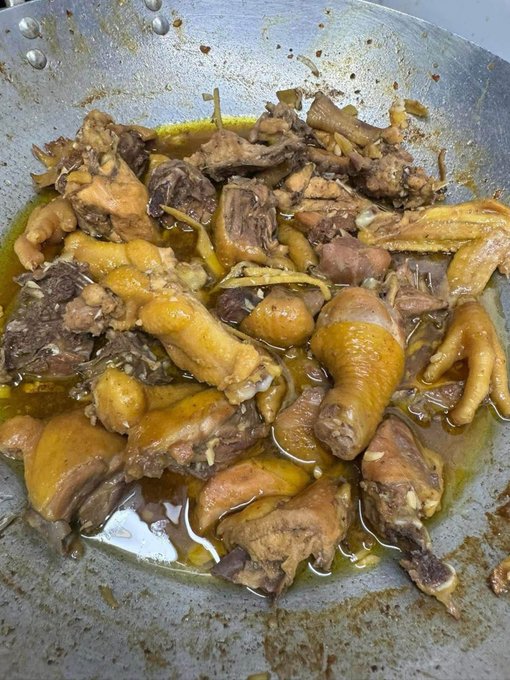 A close-up, overhead shot shows a cooked chicken dish with drumsticks and bone-in pieces braised in a savory brown sauce with ginger in a metal pan.