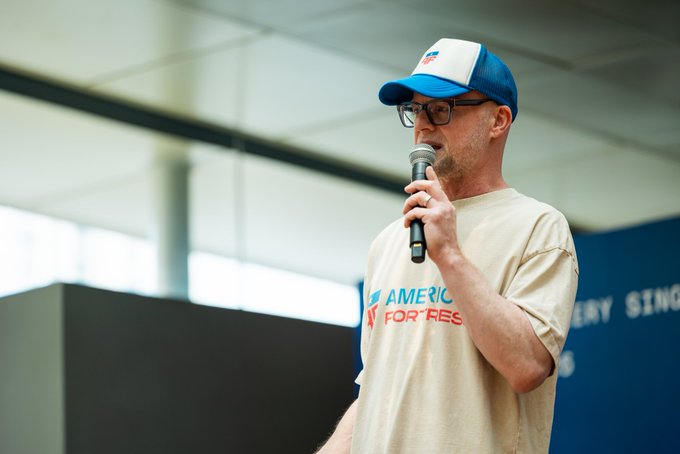 A man with glasses wearing a blue and white cap and a beige 'America for...' t-shirt speaking into a microphone at what appears to be a political or community event.