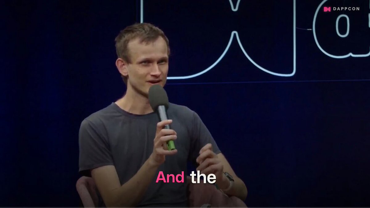 The image shows a man sitting behind a microphone, speaking into it during an event or presentation. The background of the image features a blue wall with white text that reads "And the", which appears to be part of a caption for the image. The caption also includes the names of some people who were present at the event and the name of the event itself, which is "The Ethereum Economic Zone".