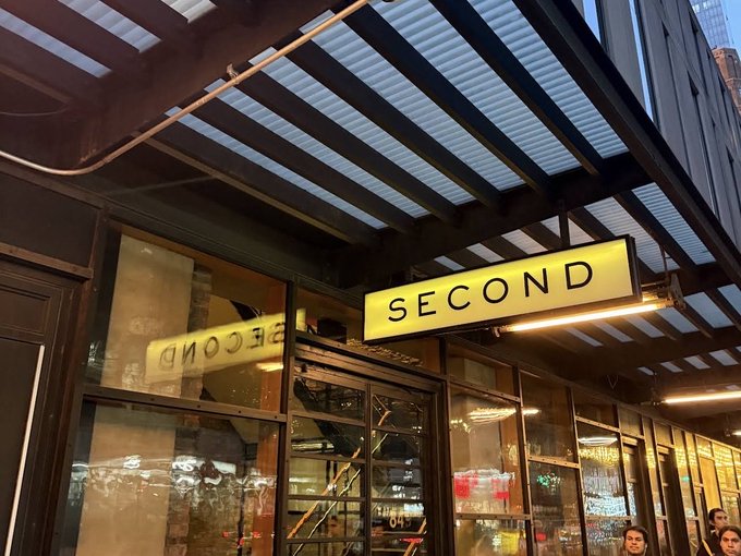 An illuminated yellow 'SECOND' sign hanging under a metal canopy at the entrance of what appears to be a bar or restaurant, with people visible in the foreground.