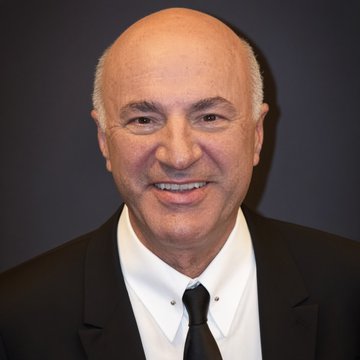 A bald man in a black suit and black tie smiles in a portrait photograph against a gray background.
