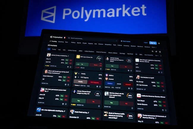 A laptop screen displays the Polymarket prediction market website, showing various political and crypto betting odds, with the Polymarket logo visible on a larger screen in the background.
