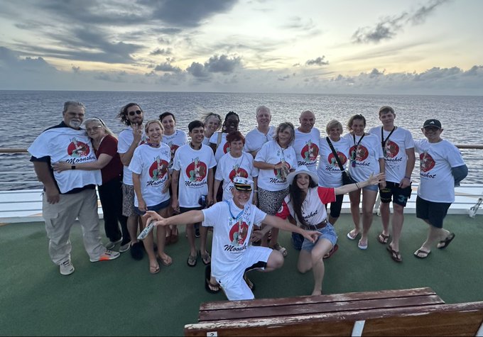 A group photo of people wearing matching white 'Moviest' t-shirts posing on a boat deck at sunset, with one person kneeling in front holding a can.