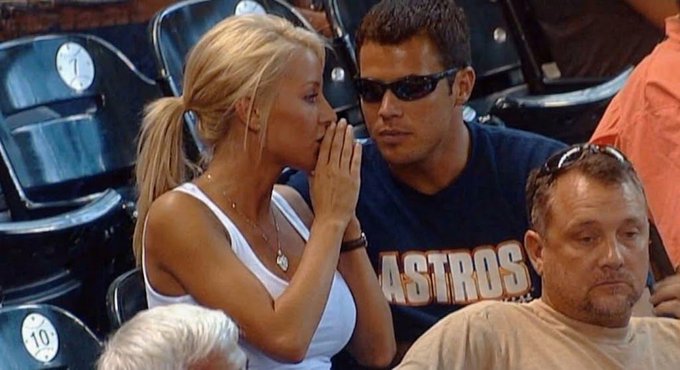 A blonde woman in a white top whispers into a man's ear while sitting in stadium seats; the man wears an 'ASTROS' shirt and sunglasses.