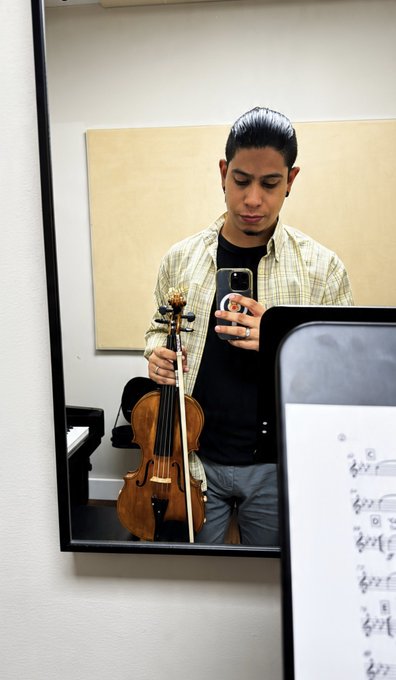 A young man with slicked-back hair takes a mirror selfie while holding a violin and bow, dressed in a yellow plaid shirt over a black t-shirt, with a music stand visible to the side.