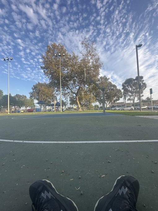 A pair of feet in black sneakers rest on a green basketball court under a vibrant blue sky dotted with wispy clouds, completing the phrase "beautiful day to" relax outdoors. A basketball hoop stands ready in the distance, with trees and parked cars lining the background.