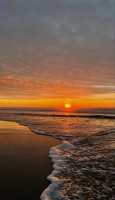 A vibrant sunset over the ocean, with the sun reflecting on wet sand and gentle waves rolling onto the shore beneath a sky filled with pink and orange clouds.