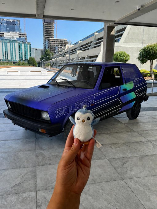 A hand holds up a small owl plushie in front of a purple car with 'SOLANA' branding parked at what appears to be a Solana event venue.