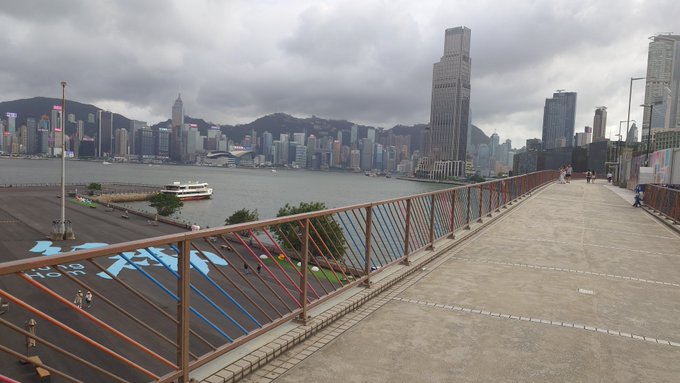 An urban skyline view of Hong Kong's Victoria Harbour with skyscrapers across the water, taken from an elevated pedestrian walkway with red and blue railings under a cloudy sky.