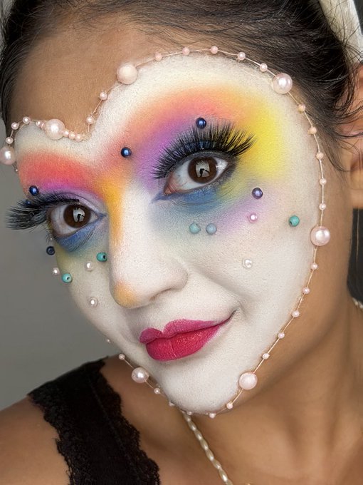 A close-up portrait of a woman with elaborate, colorful makeup featuring rainbow eyeshadow, pearls, and gems arranged on her face like a mask.