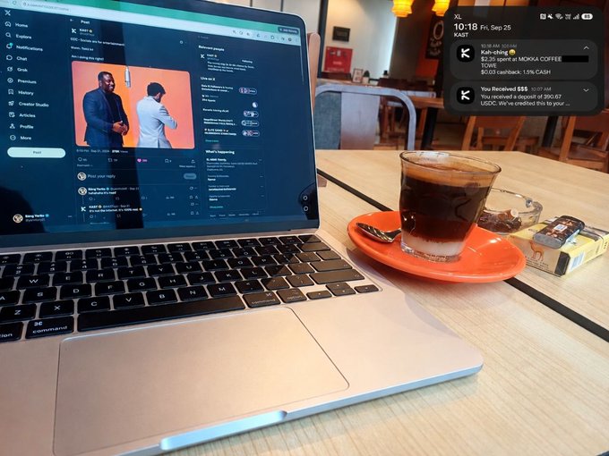 A MacBook Pro sits on a wooden cafe table displaying a social media feed, next to a glass of iced coffee on an orange saucer; a phone notification overlay in the top right shows '10:18' and messages including 'Koh-ching' and 'You Received $55' from KUCOIN.