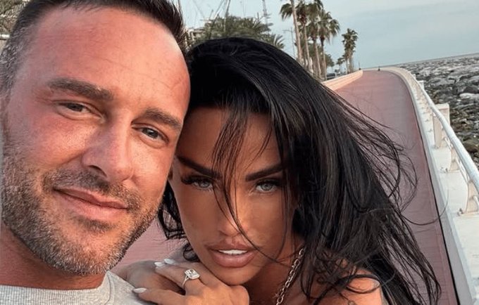 A close-up selfie of a couple outdoors with palm trees in the background; the woman has long dark hair and the man has short hair with stubble, and the woman is showing a ring on her finger.