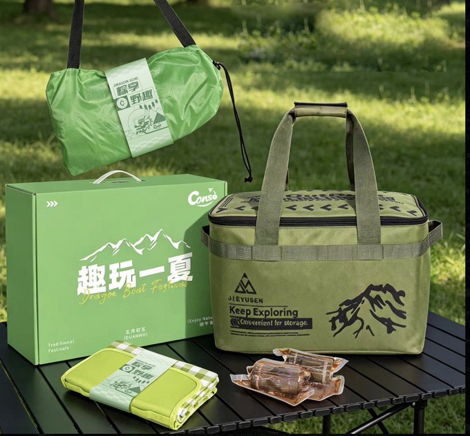 The image features a green camping bag, a green box with Chinese writing, and two bags of food sitting on a table outdoors. The scene appears to be set up for an outdoor adventure or camping trip, as indicated by the presence of these items.