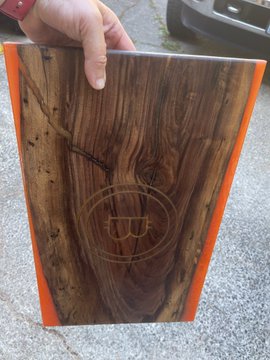 A person holds up a wooden cutting board with a decorative burned-in design in the center and orange-colored edges.