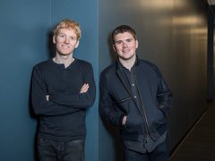 A low-resolution photograph of two men standing side-by-side against a dark blue wall; the man on the left wears a dark sweater with arms crossed, and the man on the right wears a dark jacket.