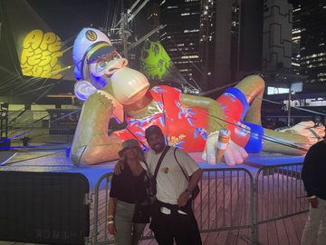 A couple poses in front of a large, brightly lit inflatable doll in a red dress, with a man appearing to vomit a green stream, set against a nighttime cityscape.