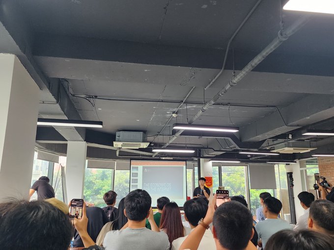 A crowded seminar or presentation room with attendees holding up phones to photograph a speaker standing by a projection screen displaying 'THE BEGINNING OF THE JOURNEY'.