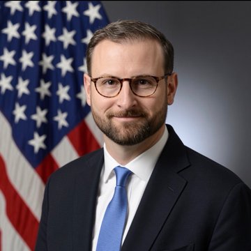 An official portrait-style photograph shows a man with a beard and glasses wearing a dark suit and blue tie, standing in front of an American flag.