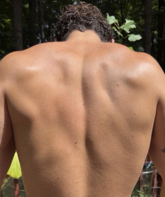 A shirtless man photographed from behind, displaying a muscular back in an outdoor setting with trees.