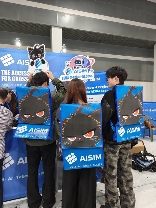Three people standing in front of an "AISIM" (eSIM, AI-Token, Access) booth, wearing promotional backpacks featuring a black cat design and holding up cat-themed signs.