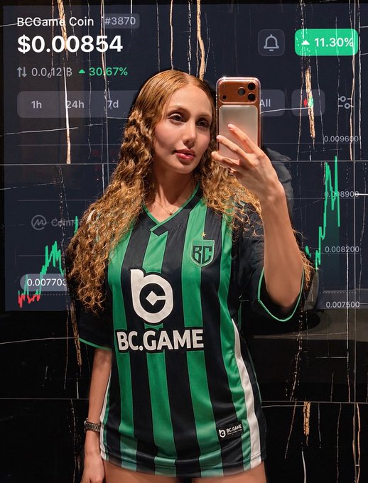 The image features a woman taking a selfie with her cell phone, wearing a green and black striped soccer jersey. The background of the photo includes a computer screen displaying stock market data, which seems to be related to the BC Game. This suggests that the woman might have been discussing or planning for sports betting on the BC Game while taking the selfie.