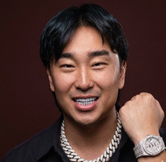 A smiling man with braces, wearing a black shirt and diamond-encrusted jewelry including a watch and chains, poses against a dark red background.