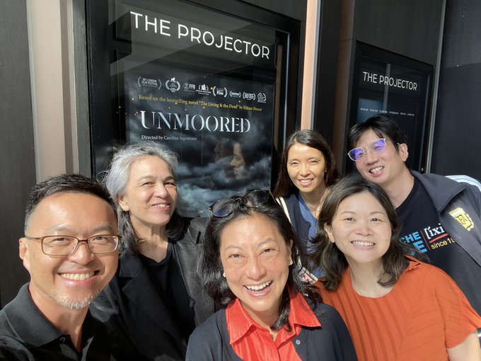 A group of six smiling people pose for a photo in front of 'THE PROJECTOR' cinema, where the film 'UNMOORED' directed by Caroline Ingvarsson is being shown.
