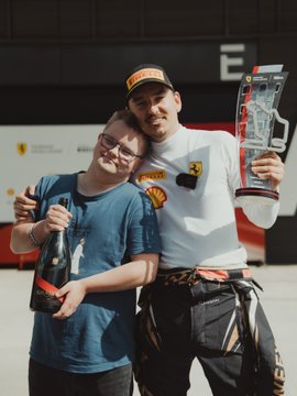 A man and woman pose together at what appears to be a racing event; the man wears a Ferrari cap and racing suit while holding a small trophy, and the woman smiles beside him.