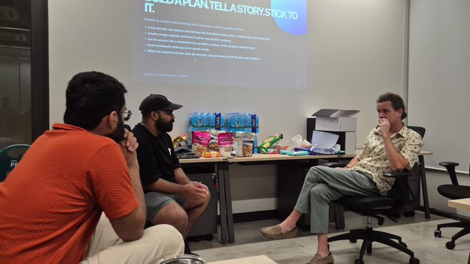 Three men are seated in a meeting room with a presentation projected behind them, while a table of snacks and water bottles sits nearby.