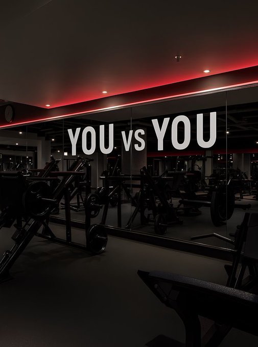 A dimly lit gym interior with red accent lighting along the ceiling, featuring weightlifting equipment and a large mirror displaying the motivational text 'YOU vs YOU'.