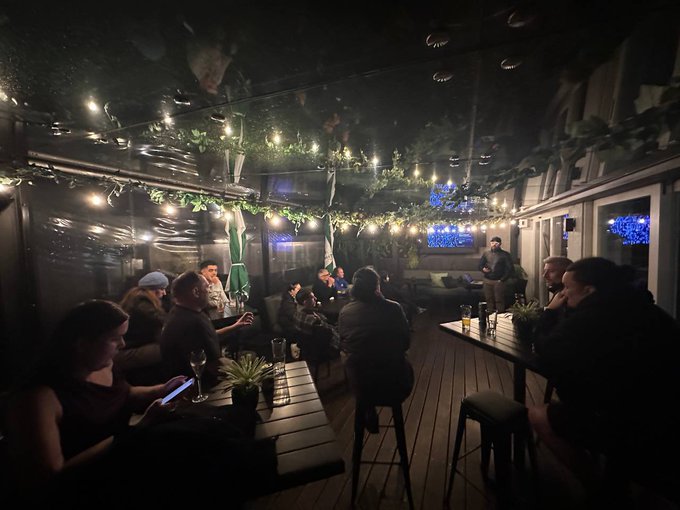 A dimly lit, atmospheric bar or lounge area decorated with hanging string lights and artificial vines, where several patrons are seated at tables.