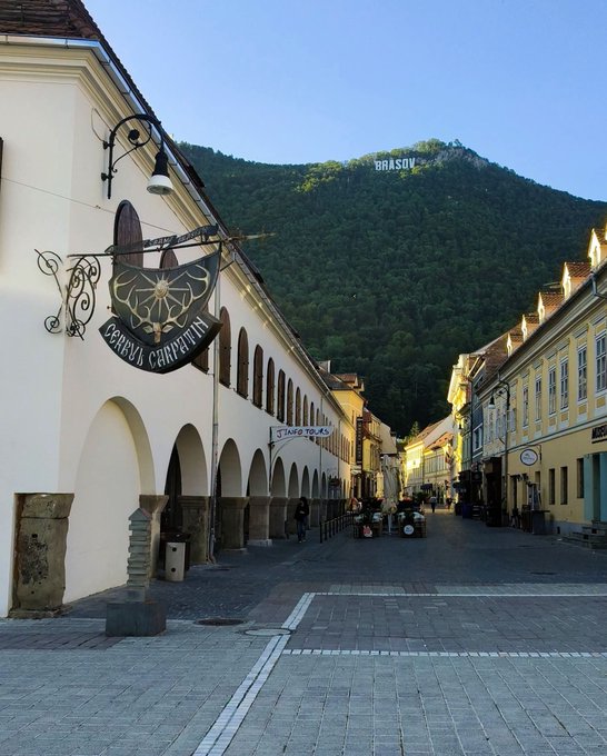A street view in Brașov, Romania, showing the historic 'Cerbul Carpatin' building with arched colonnades, with the 'BRAȘOV' sign visible on Tâmpa mountain in the background.