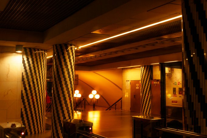 A dimly lit, empty subway or underground station features distinctive geometric-patterned columns in yellow and black, with warm amber lighting illuminating the polished floor and tiled walls. The image serves as the "Transition" limited edition artwork (1 of 5) listed for 4 XTZ on objkt.com, evoking a sense of fleeting passage through an abandoned, atmospheric transit space.