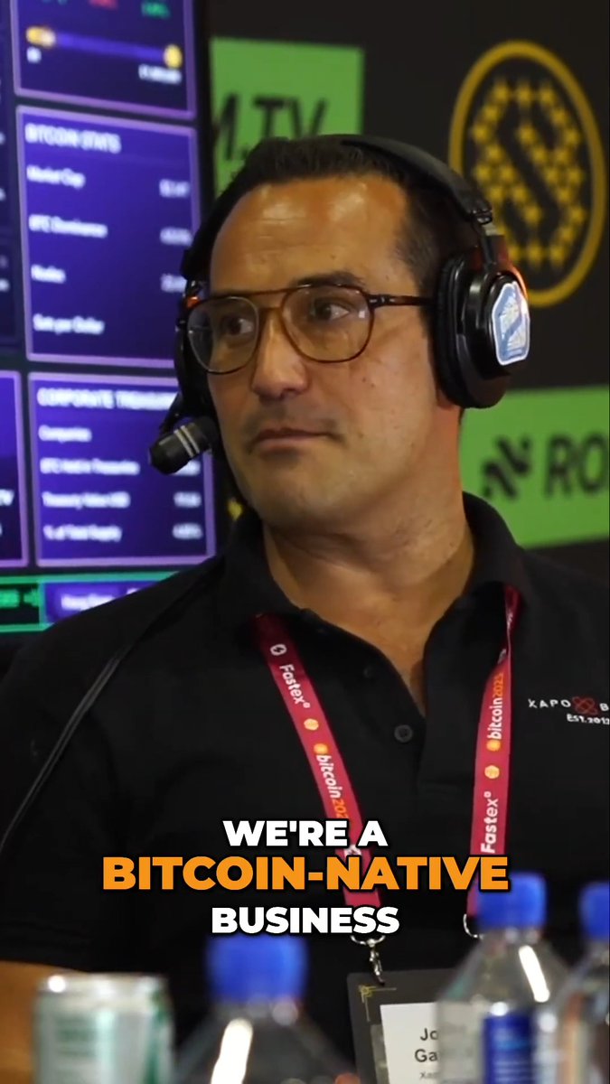 A man with glasses and a headset speaks at a conference with 'Bitcoin 2025' branding, with the caption 'WE'RE A BITCOIN-NATIVE BUSINESS' overlaid in white and orange text.