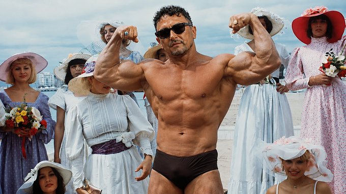 A muscular man in sunglasses and small black swim briefs flexing in front of a group of women wearing vintage dresses and sun hats.