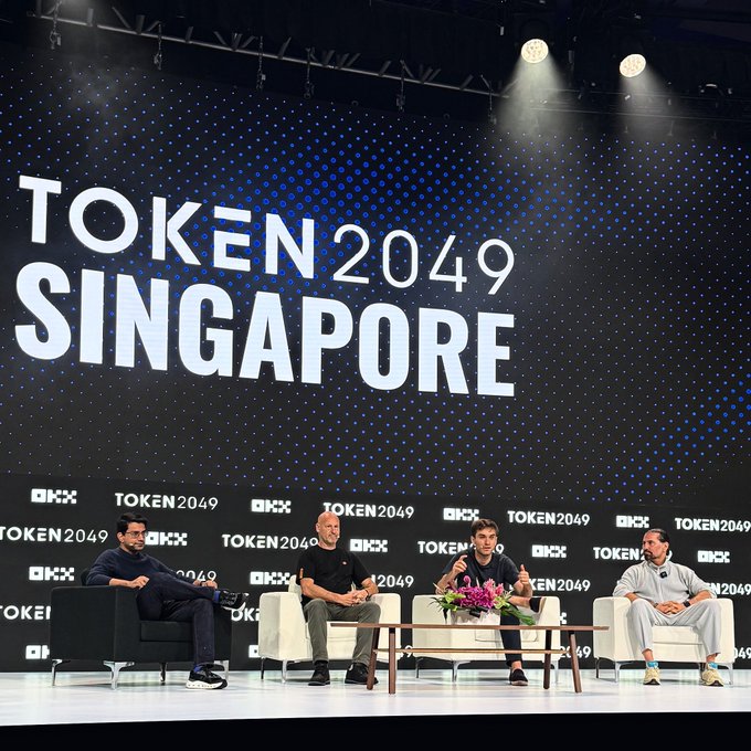 A photograph of a panel discussion at the 'TOKEN 2049 SINGAPORE' event, showing four men seated on stage in front of a large black backdrop with the event's logo and sponsor 'OKX'.