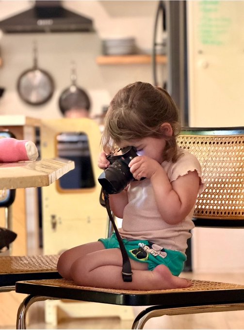 A young child sits on a rattan chair in a kitchen setting, looking intently through a DSLR camera held up to their face.