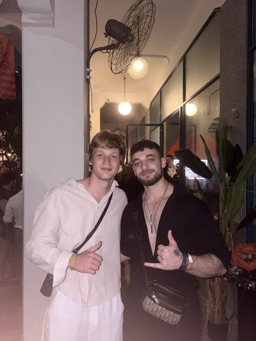 Two young men pose together indoors, one in a white striped shirt and the other in a black open shirt with a crossbody bag, both making hand gestures toward the camera.