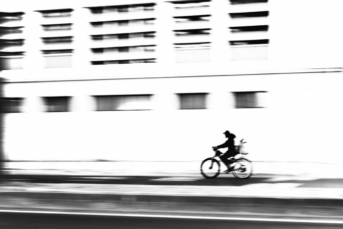 A black-and-white motion-blurred photograph capturing a cyclist riding past a building, with the panning technique creating a dynamic sense of movement and speed.