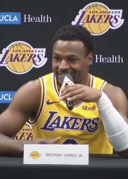 Bronny James Jr. sits at a press conference table wearing a Lakers jersey, smiling while biting on the Lakers logo, with a nameplate reading 'BRONNY JAMES JR.' in front of him.