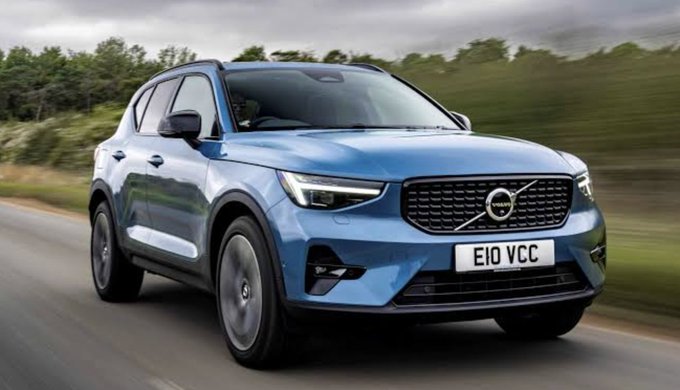 A blue Volvo SUV (license plate "E10 VCC") is shown driving on a road, with the article noting Volvo's development of a proprietary cryptocurrency using Cardano's blockchain to test transactions with material and transport suppliers. The vehicle represents the automotive giant leveraging blockchain technology in its supply chain operations.