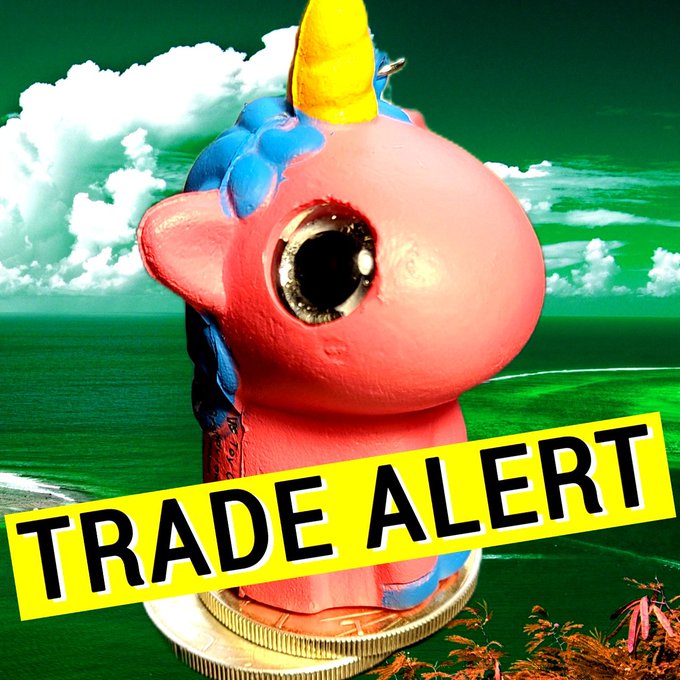 A pink and blue unicorn figurine sitting on a coin against a green cloudy sky, overlaid with a yellow 'TRADE ALERT' banner.