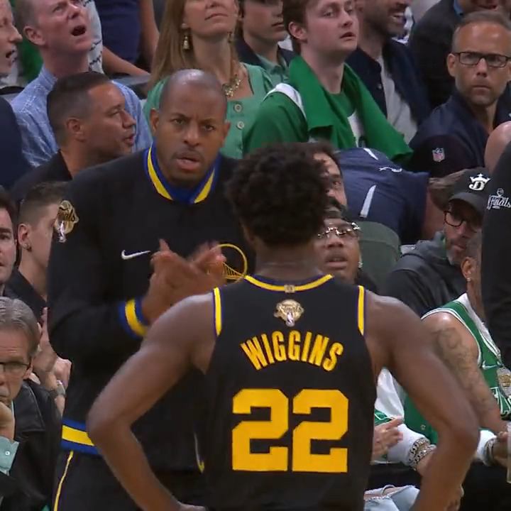 Photo from an NBA Finals game showing Golden State Warriors player Wiggins (#22) talking with a coach on the bench, with fans in the background including some wearing Boston Celtics gear.